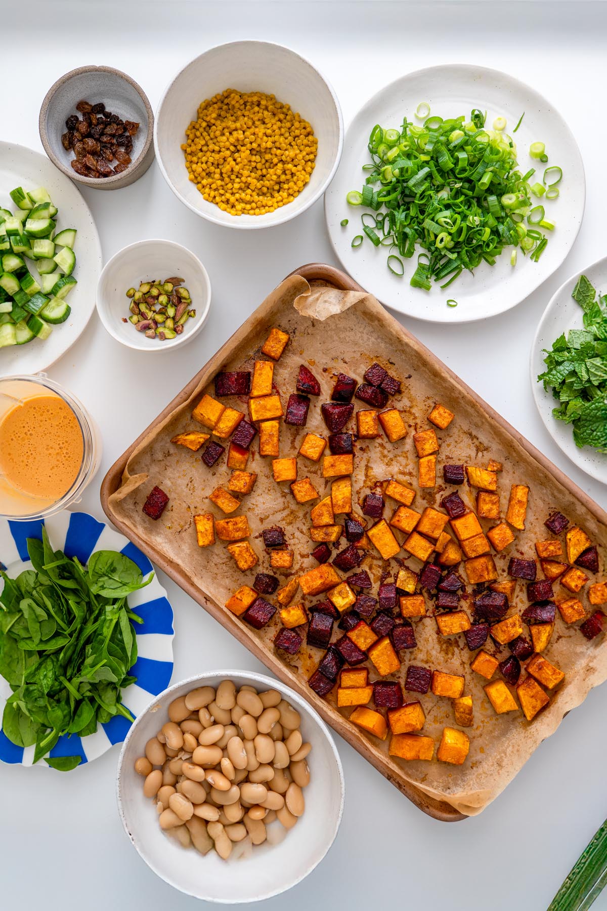 Colorful fresh ingredients for Moroccan Spiced Pearl Couscous Salad arranged around a baking tray on a white surface.