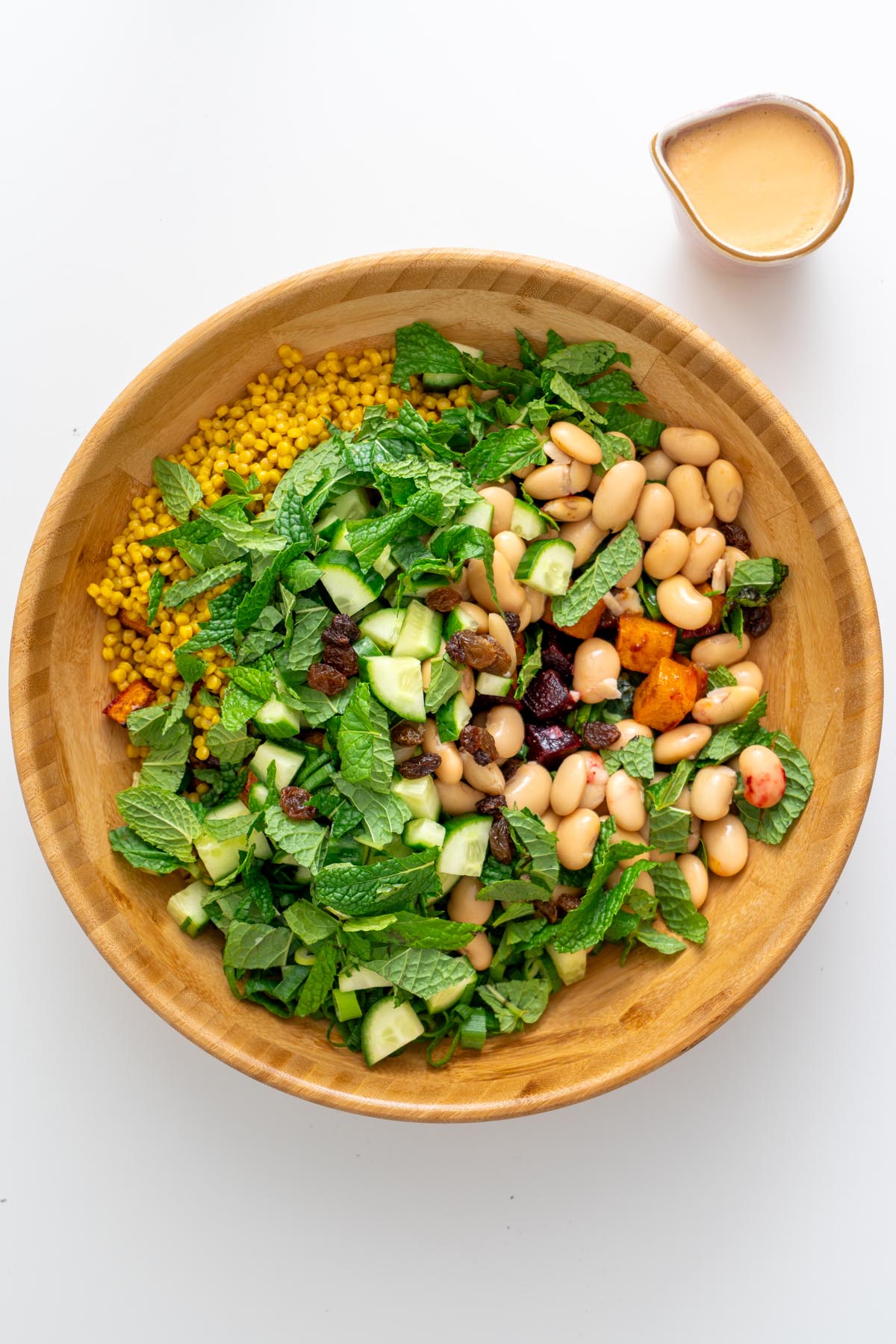 Unmixed ingredients for Moroccan Spiced Pearl Couscous Salad arranged in a wooden bowl with tahini dressing on the side.