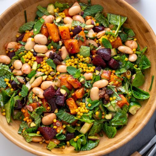Overhead view of Moroccan Spiced Pearl Couscous Salad in a wooden bowl next to a cup of tahini dressing and mint leaves.
