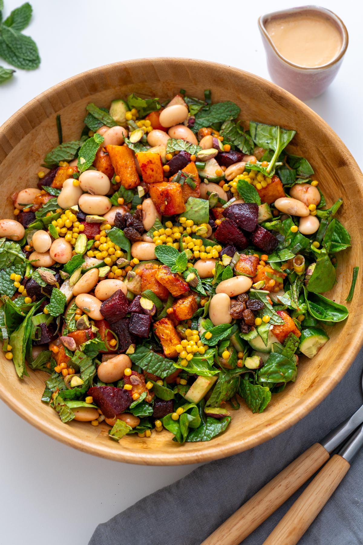 Overhead view of Moroccan Spiced Pearl Couscous Salad in a wooden bowl next to a cup of tahini dressing and mint leaves.
