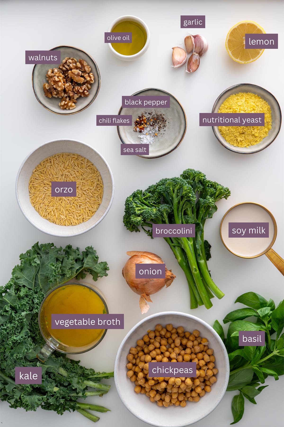 Vibrant ingredients for a creamy pesto orzo on a white surface, including kale, basil, chickpeas, broccolini, and nutritional yeast