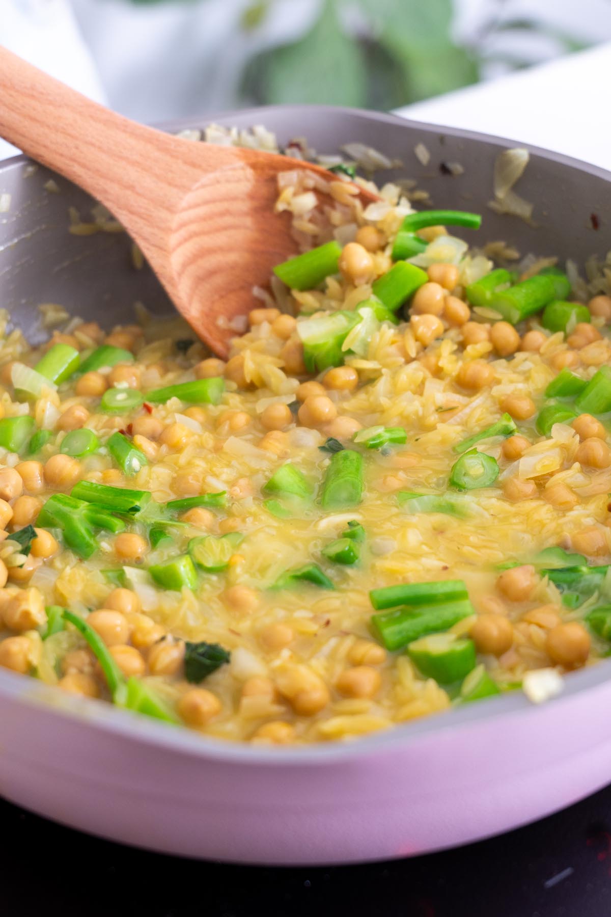Creamy orzo cooking in a skillet with chickpeas and chopped broccolini, being stirred with a wooden spoon