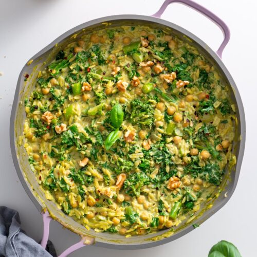 Finished creamy pesto orzo in a pink skillet with wilted kale, chickpeas, broccolini, and walnut garnish