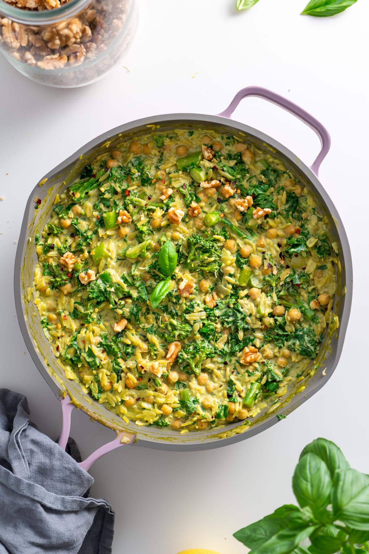 Finished creamy pesto orzo in a pink skillet with wilted kale, chickpeas, broccolini, and walnut garnish