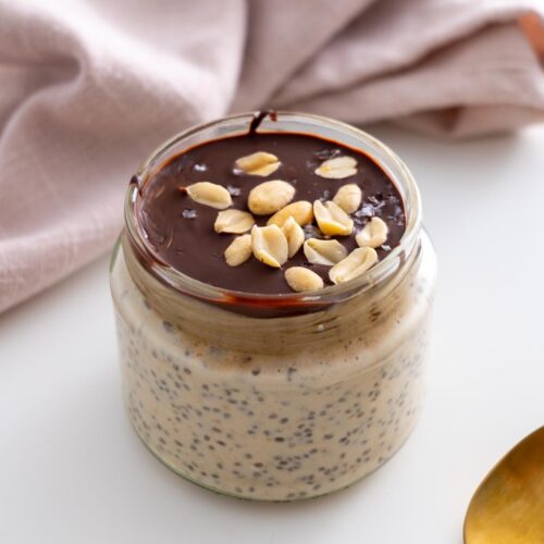 Snickers overnight oats in a jar topped with melted chocolate and peanuts, styled with a gold spoon and pink napkin.