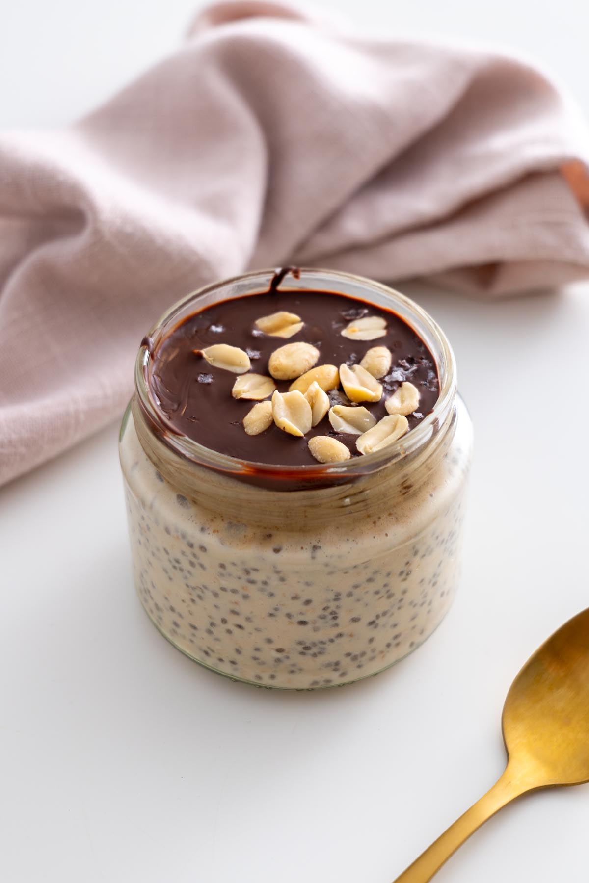 Snickers overnight oats in a jar topped with melted chocolate and peanuts, styled with a gold spoon and pink napkin.