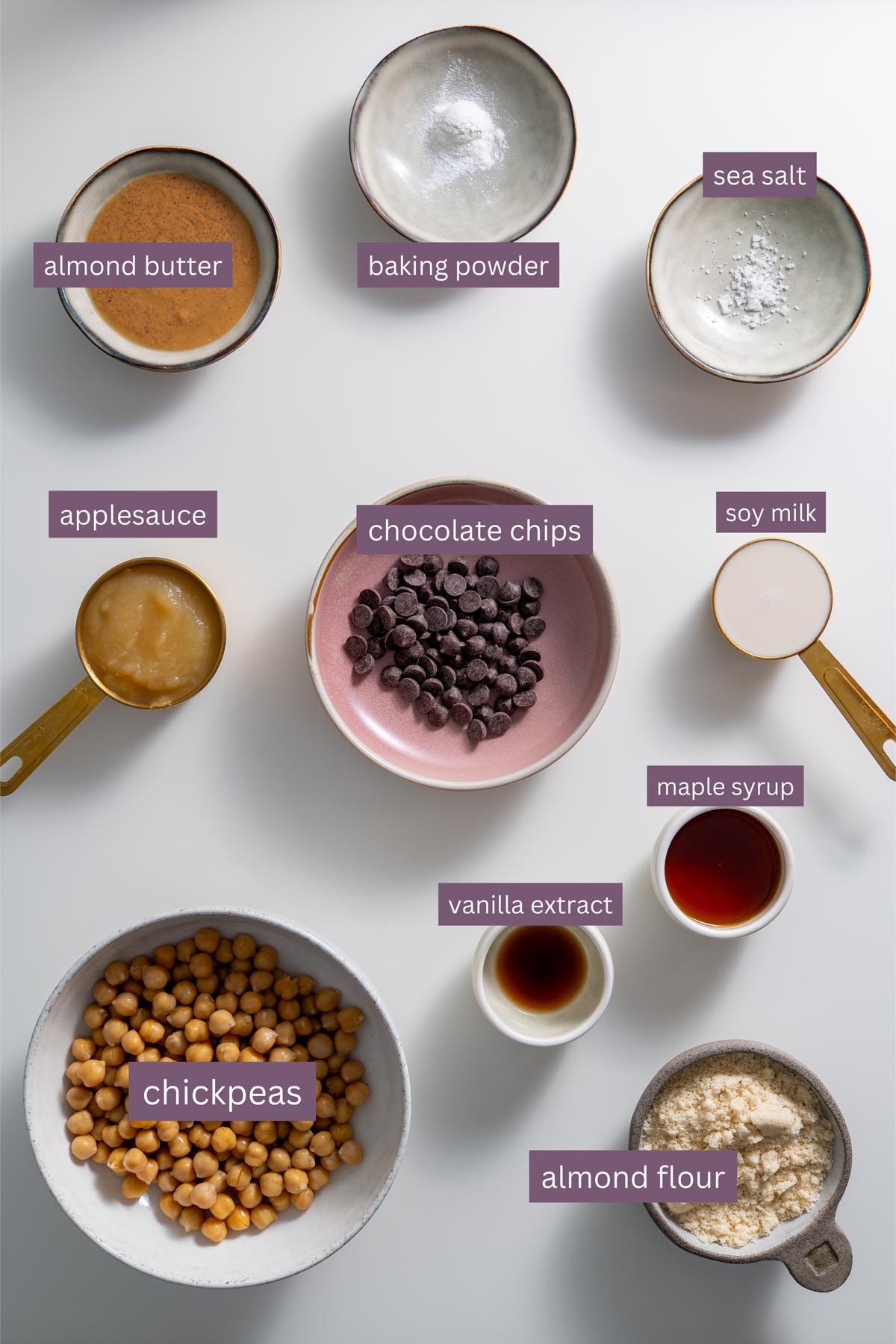 Flat lay of labeled ingredients for chickpea cookie bowls including almond butter, applesauce, almond flour, baking powder, maple syrup, soy milk, and vanilla extract.