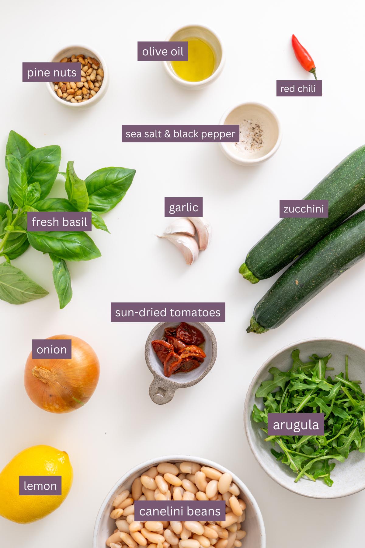 Main ingredients for zucchini bean recipe including zucchini, cannellini beans, basil, lemon, garlic, onion, chili, sun-dried tomatoes, arugula, pine nuts, olive oil, and seasonings on a white surface
